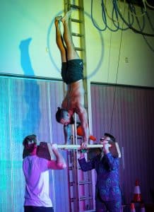 man doing handstand in air, balancing on bars which sit on the shoulders of two other men.