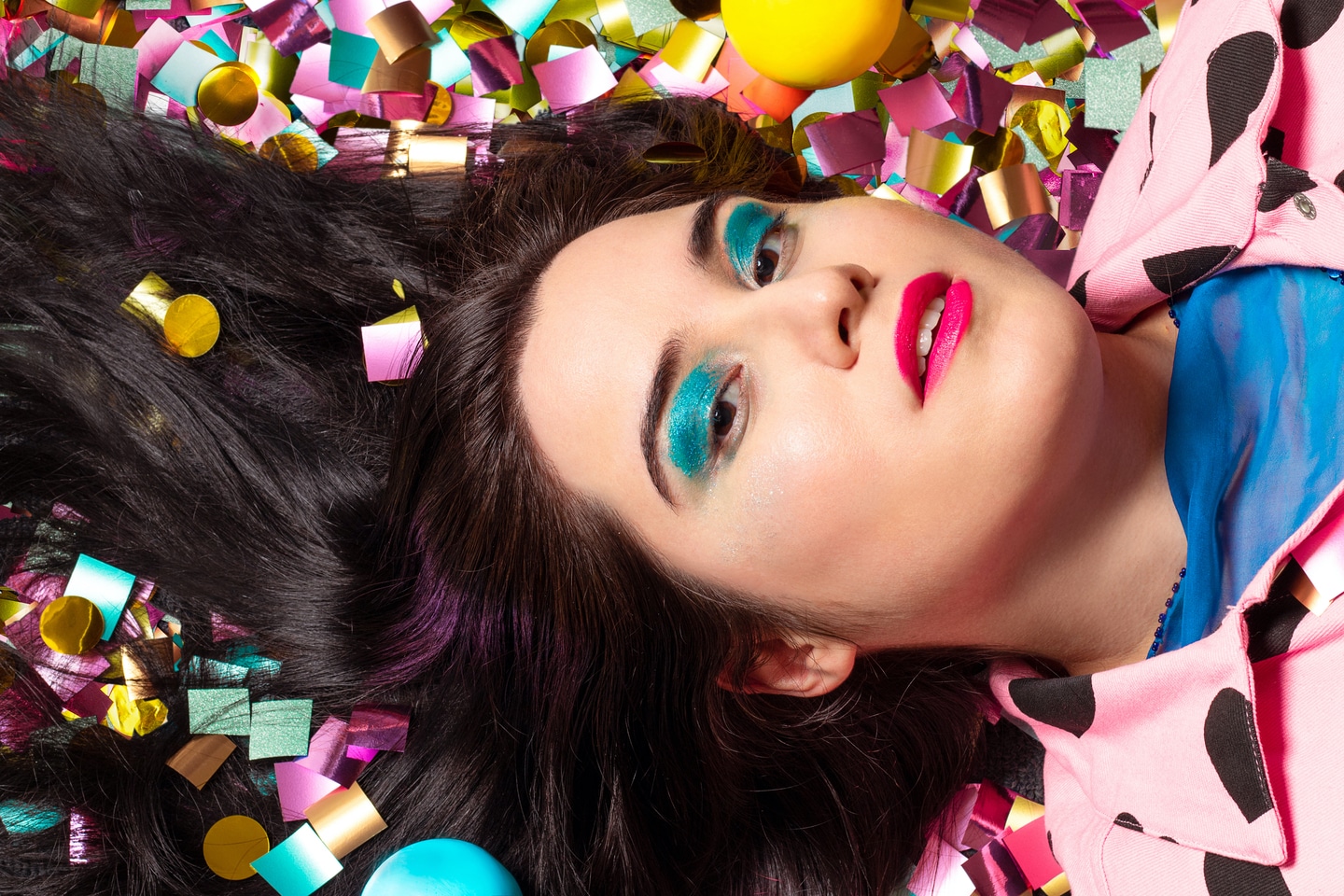 A woman with long brown hair, brown eyes with blue eyeshadow on a bed of coloured confetti