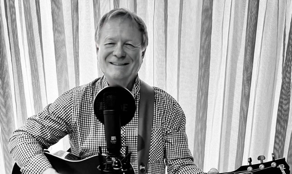 Michael G Jones Hero Image - Anywhere Festivals Smiling man in a checkered shirt plays an acoustic guitar and sings into a microphone in a home studio setup.