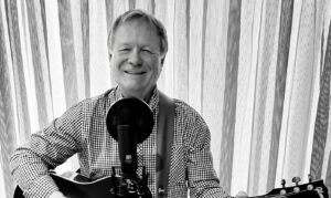 Smiling man in a checkered shirt plays an acoustic guitar and sings into a microphone in a home studio setup.