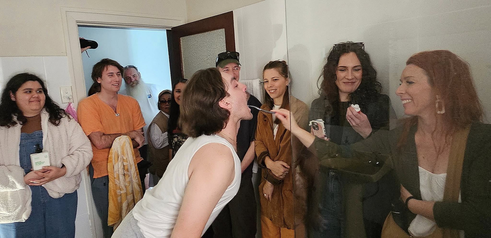 A group of friends indoors, smiling and watching as one person leans in toward another’s open mouth while a long object is held near him, suggesting a playful activity or demonstration.