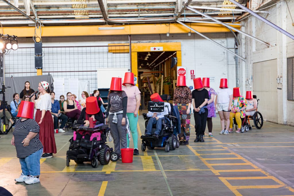 The opening line of performers wearing red buckets - a scene from Screech Arts’'The Bucket list' at the 2026 Anywhere Festival (Image Credit: Creative Futures Photography).