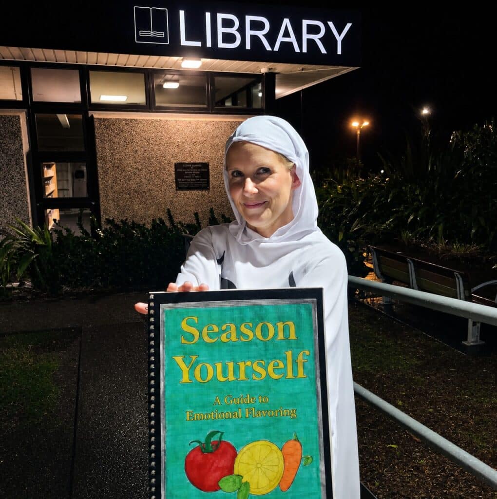 Phoebe Maher, in a ghost costume,  stands with her book in front of a sign that says "Library".