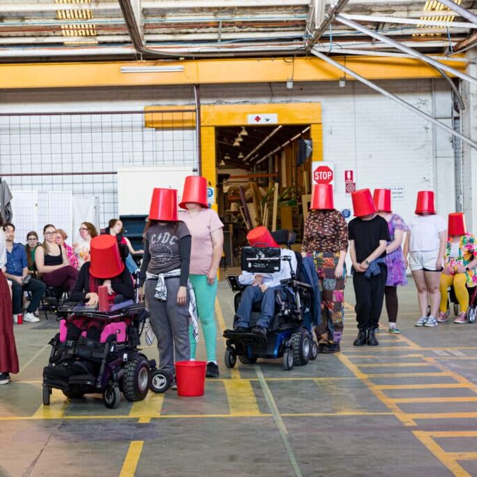 a line of people wearing red buckets on their heads, during Screech Arts’ latest show, The Bucket List.