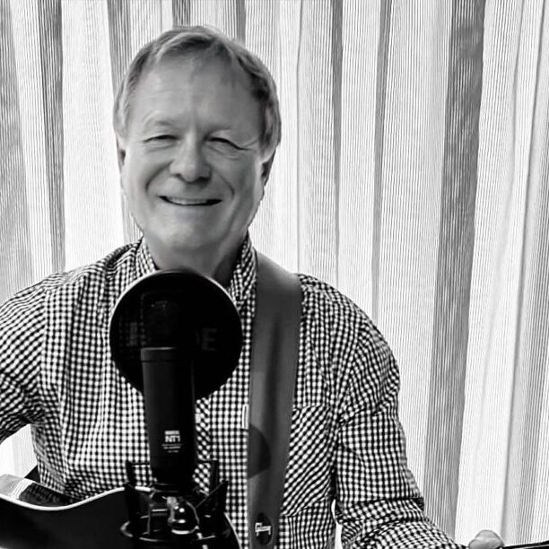 Smiling man in a checkered shirt plays an acoustic guitar and sings into a microphone in a home studio setup.